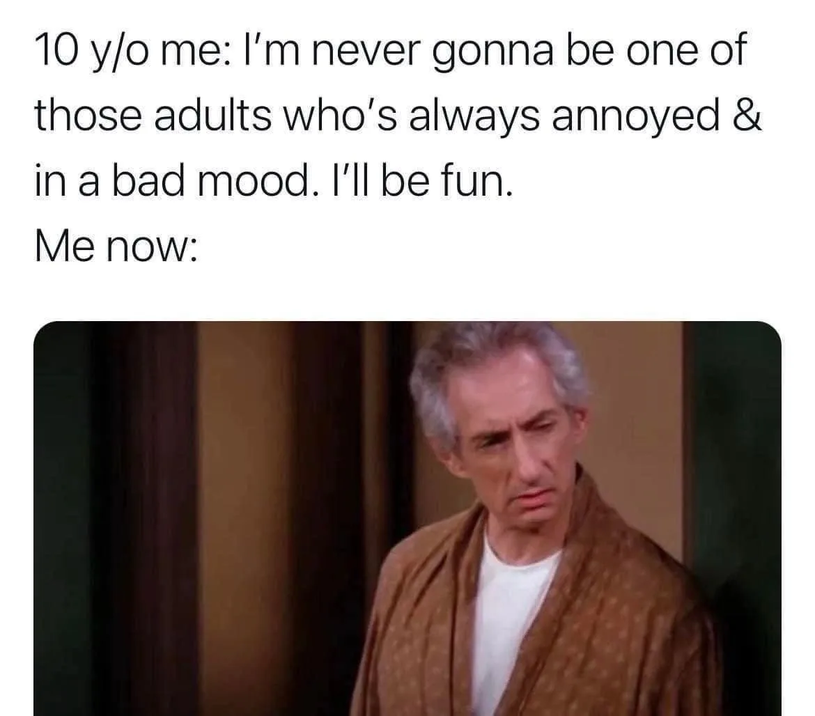The image shows a man with gray hair wearing a brown robe over a white t-shirt, looking down with a tired, exasperated…