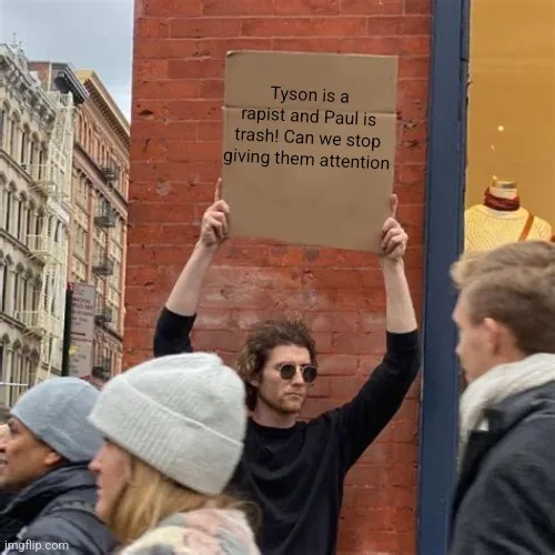 A man in sunglasses holds a cardboard sign with text accusing Tyson and Paul of being rapists and trash, urging people to…
