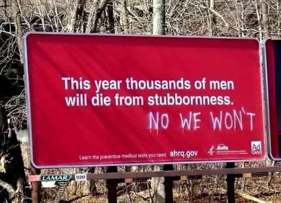A red public service announcement billboard in a wooded area reads 'This year thousands of men will die from stubbornness.'…