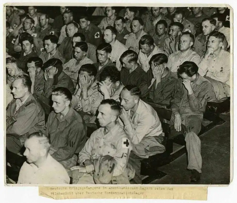 A black-and-white historical photograph shows rows of German prisoners of war seated in a lecture hall or auditorium, many…
