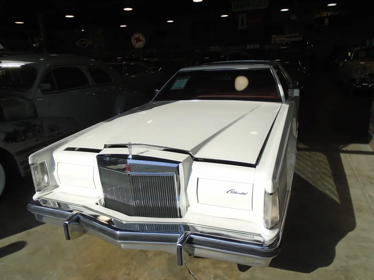 A white 1970s Lincoln Continental is displayed in a dimly lit car showroom, with other vintage cars visible in the…
