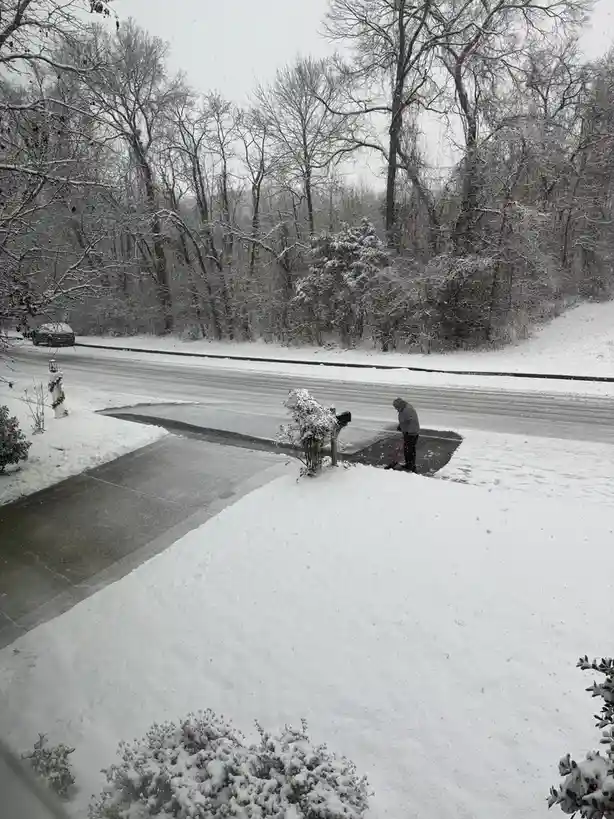 A person is shoveling snow off a driveway in a snowy residential area with bare trees and snow-covered bushes. The overlaid…