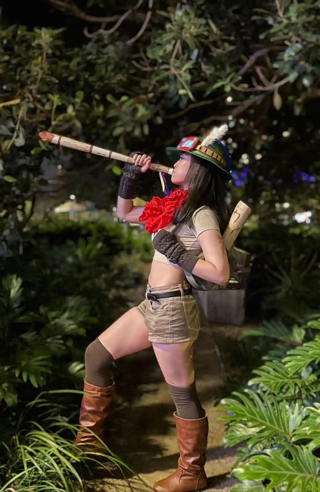 A person in a jungle explorer costume, complete with a green hat with a feather, red scarf, and brown boots, stands in a…