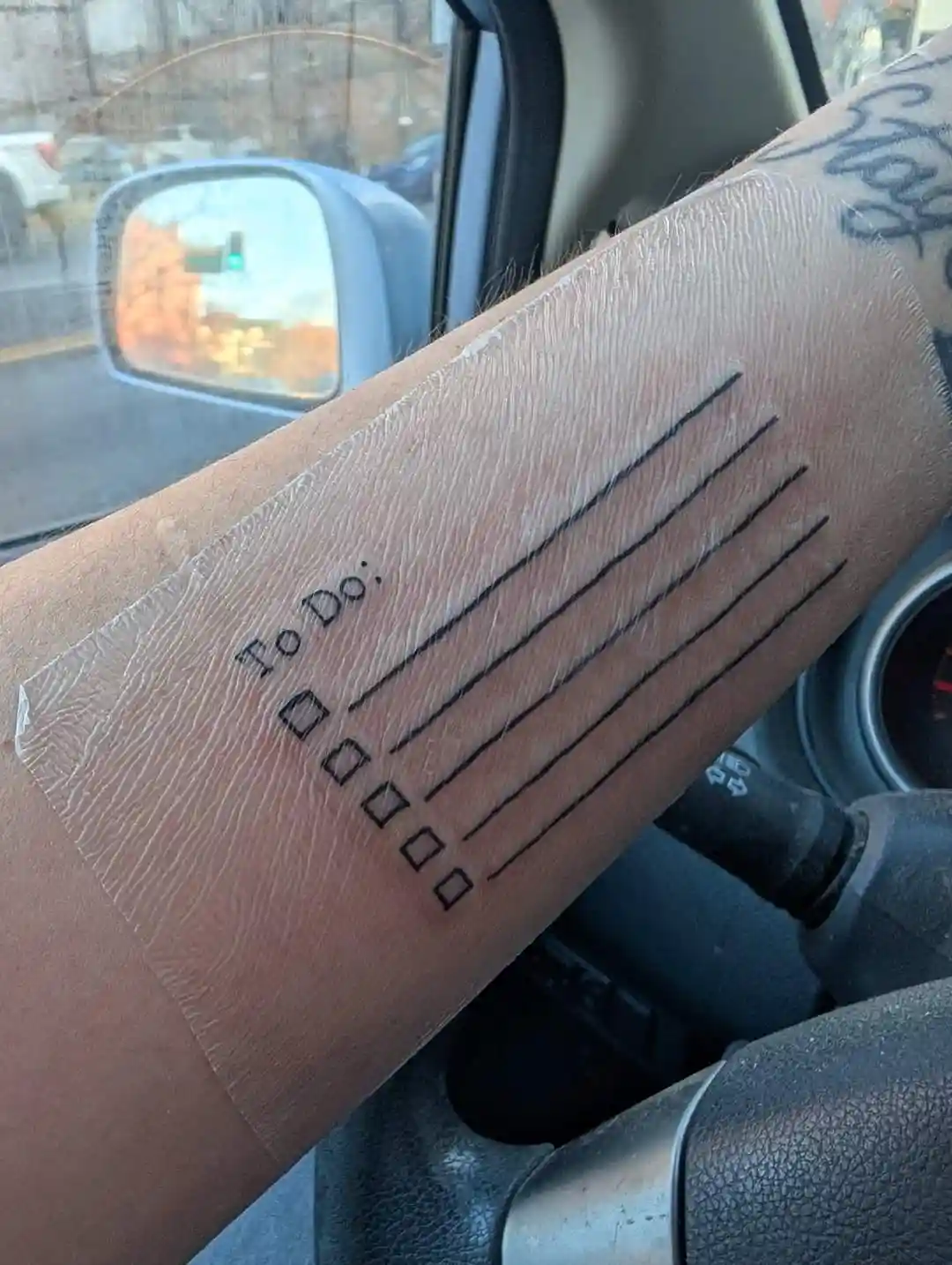 A person's forearm inside a car has a temporary tattoo of a to-do list with the heading 'To Do:' followed by four blank…
