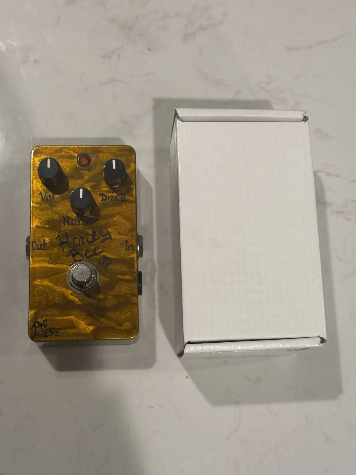 A golden guitar effects pedal with handwritten text reading 'Honey Bee' sits next to a plain white box on a light surface…