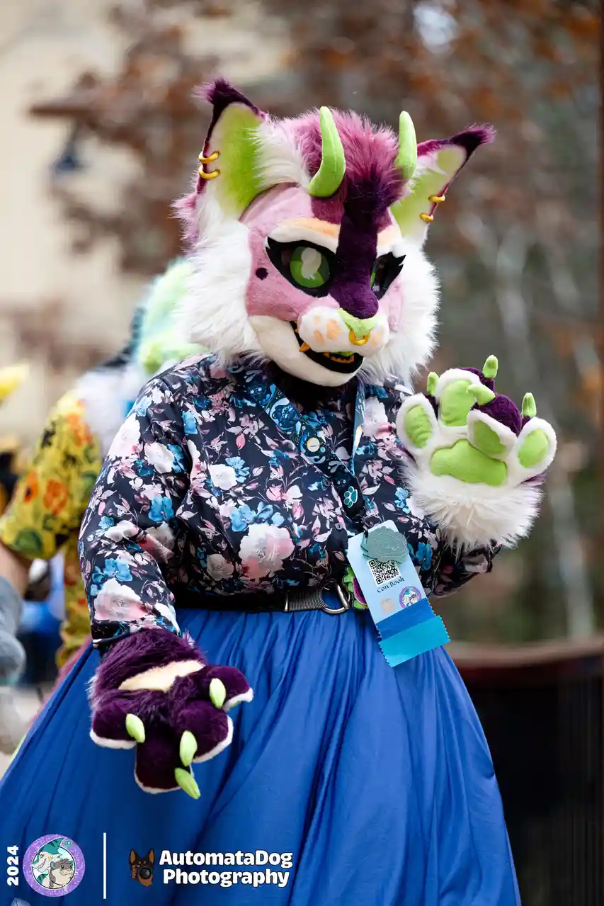 A person in a detailed pink, purple, and green fursuit with horns and large ears wears a floral shirt and blue skirt…