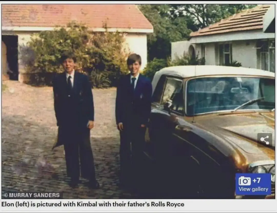 Two young boys in dark suits stand beside a vintage Rolls Royce in a driveway. The boy on the left is identified as Elon…