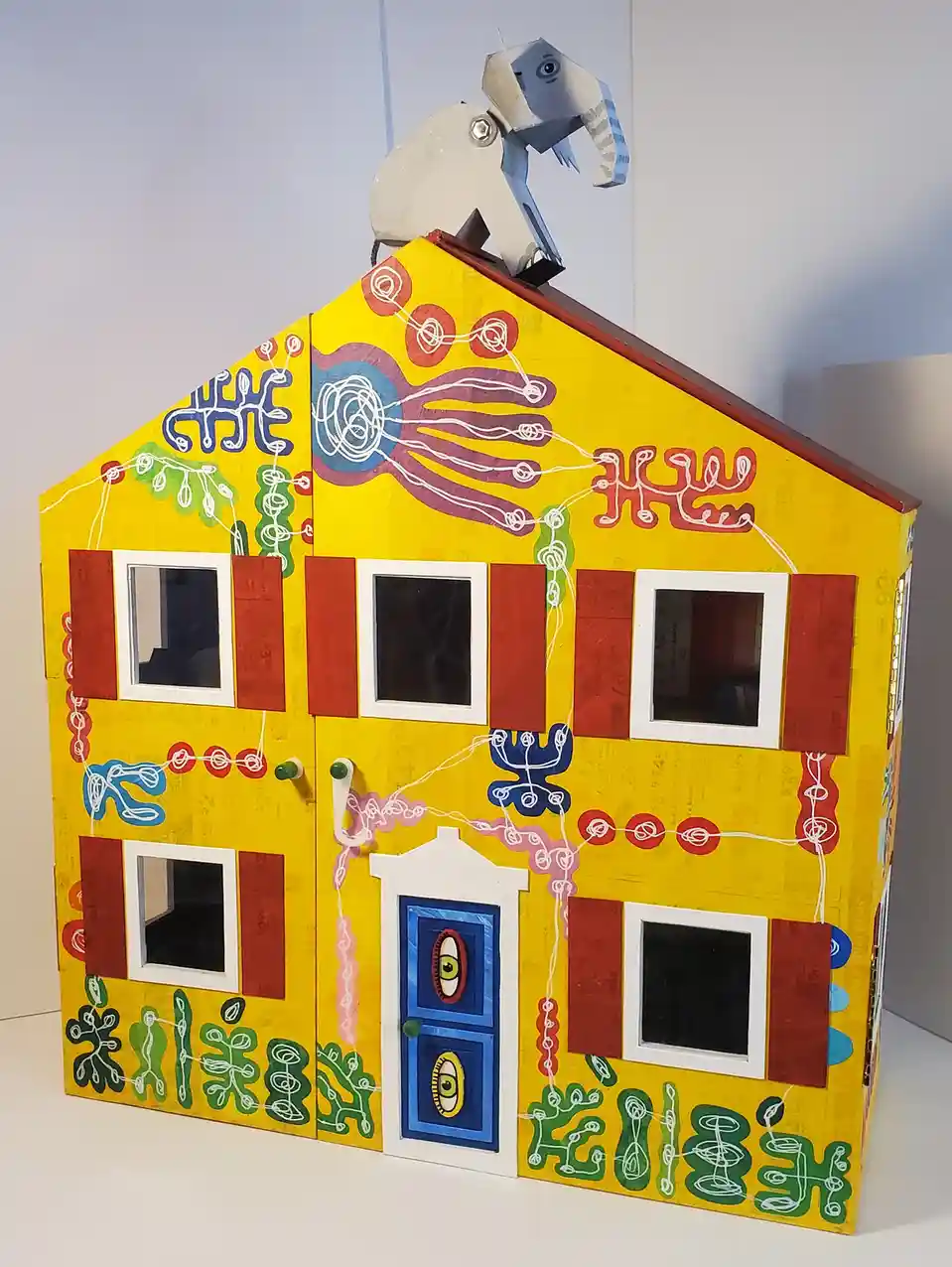 a brightly colored yellow cardboard house with red shutters, windows, and a blue door featuring two cartoon eyes. White…