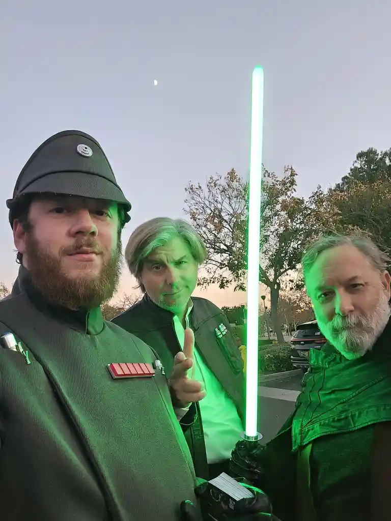 Three men dressed in Star Wars costumes pose outdoors at dusk, one holding a glowing green lightsaber. The man on the left…