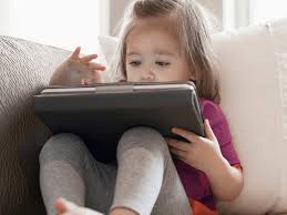 A young child with light brown hair sits on a couch, intently focused on a tablet computer held in her lap. She wears a…