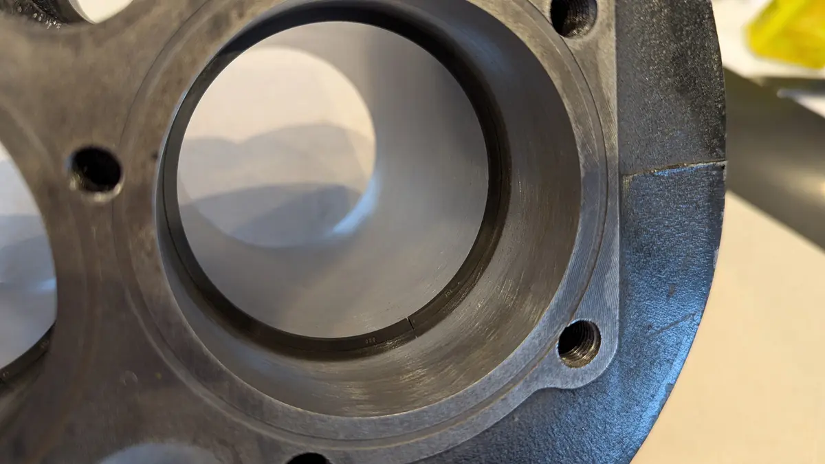 A close-up photo of a metal mechanical part with a large central bore and several bolt holes around its circumference. The…