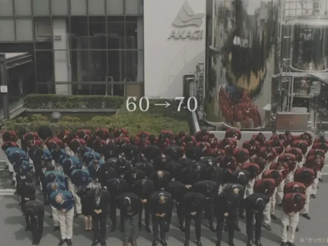 A large group of people in business attire are bowing deeply in unison outside a building with 'AKACI' signage. White text…