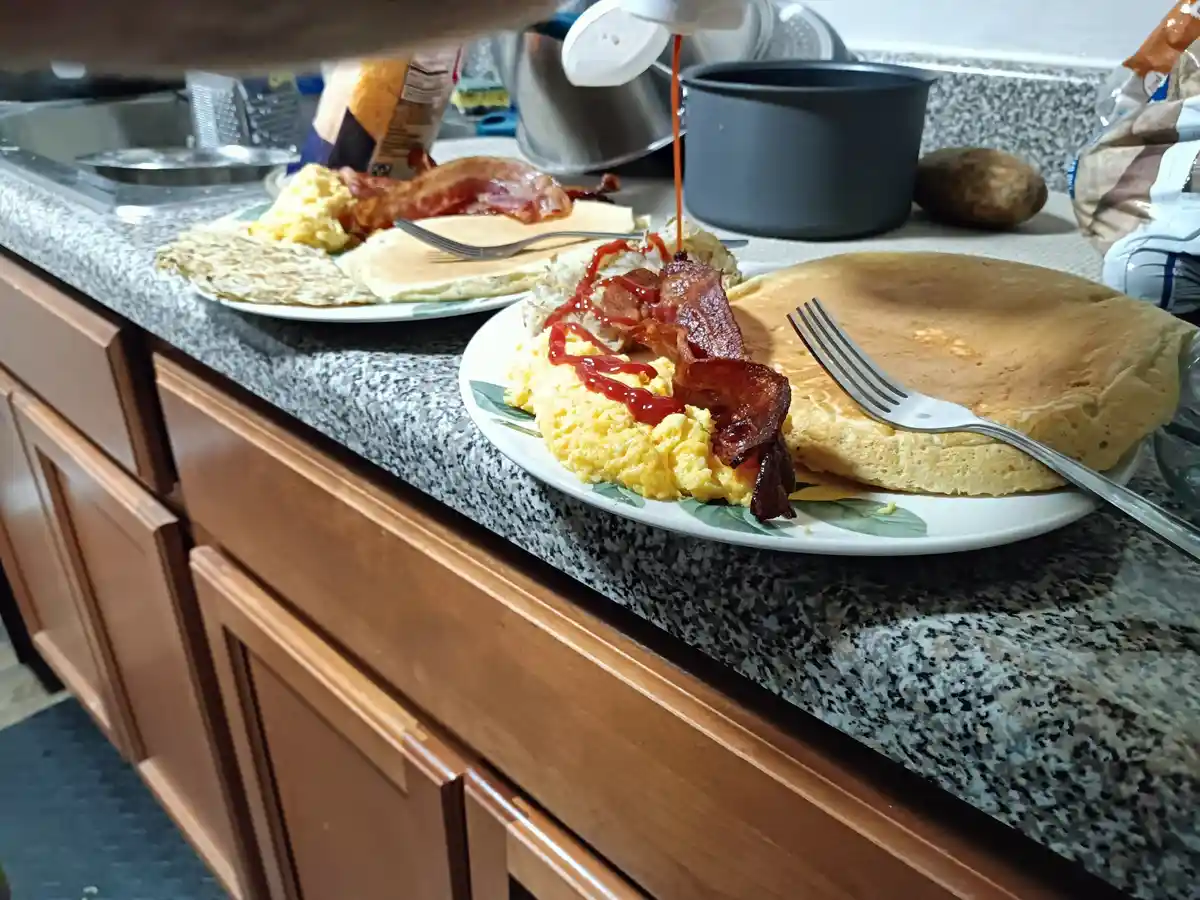 A kitchen counter holds two plates of breakfast food, including scrambled eggs, bacon, and pancakes, with ketchup being…