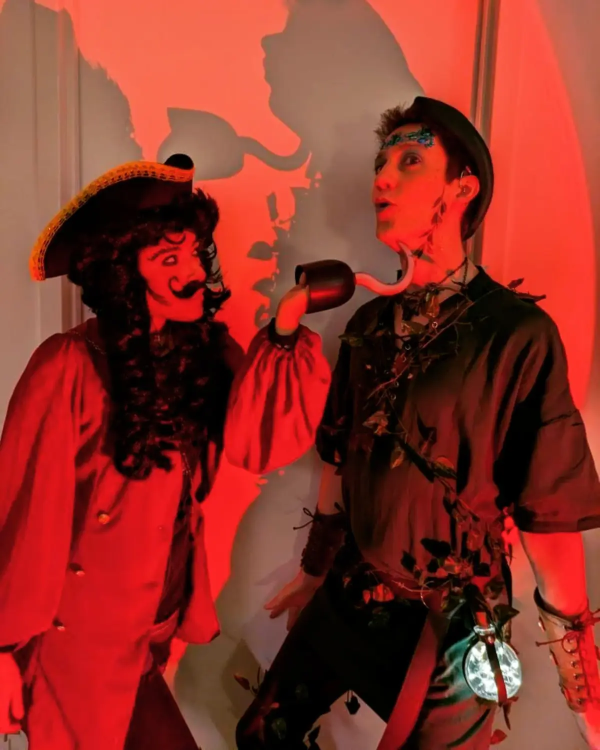 Two people in elaborate pirate-themed costumes stand under red lighting. The person on the left wears a tricorn hat and has…