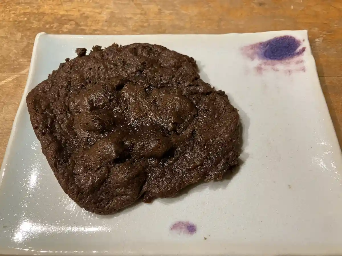 A close-up photo shows a large, dark brown, lumpy baked good on a white square plate, which has purple and pink stains on…