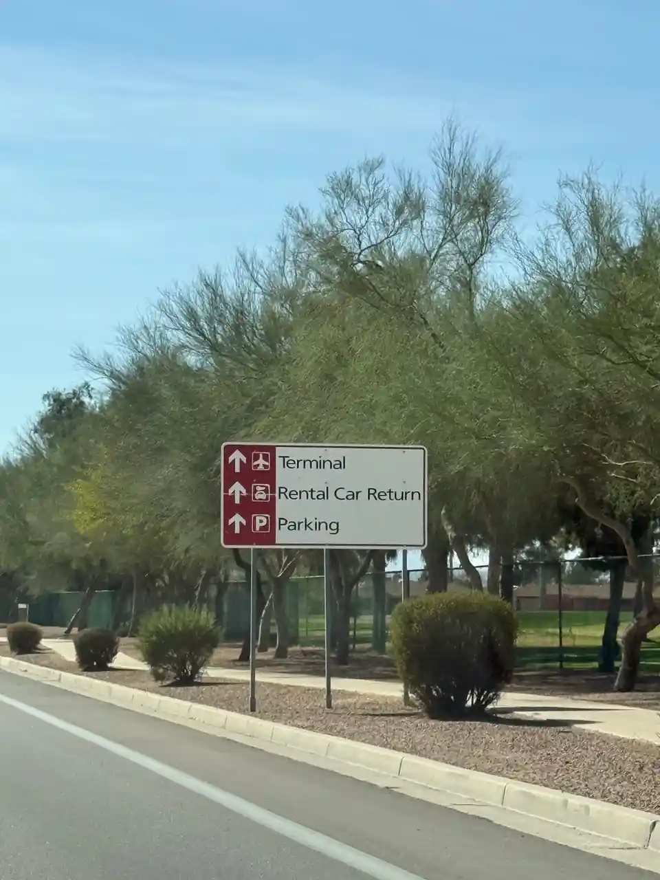 A roadside sign at an airport directs travelers to Terminal, Rental Car Return, and Parking, all with arrows pointing…