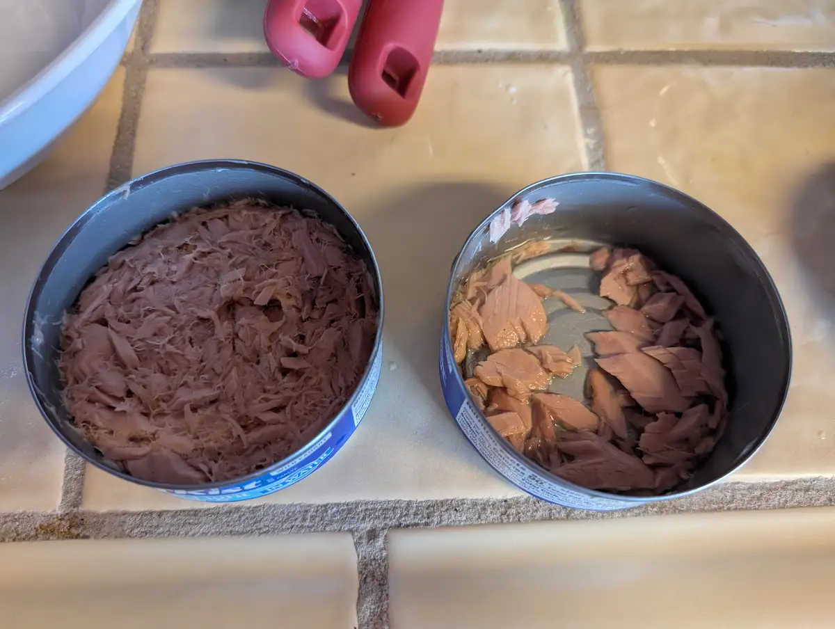 Two open tuna cans sit side by side on a tiled kitchen counter; one contains finely flaked tuna in water, the other has…