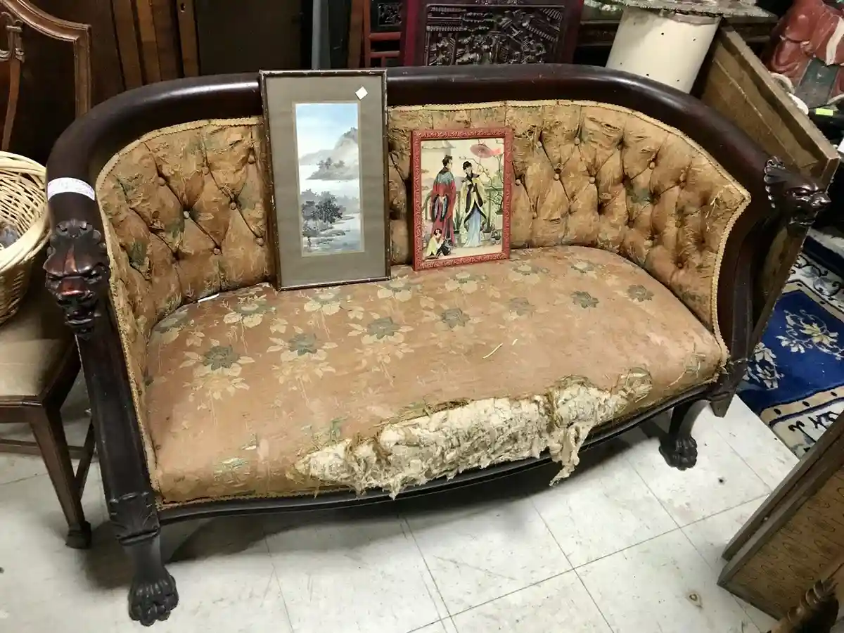 An old, worn-out tufted sofa with floral fabric and dark wood frame has a large, frayed tear in its seat cushion. Two framed…