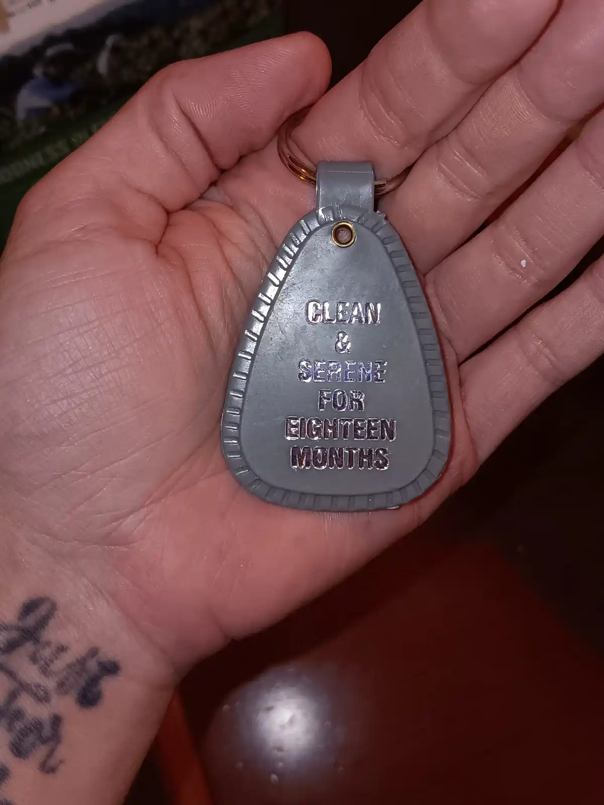 A person holds a gray, teardrop-shaped keychain tag with raised lettering that reads 'CLEAN & SERENE FOR EIGHTEEN MONTHS'…
