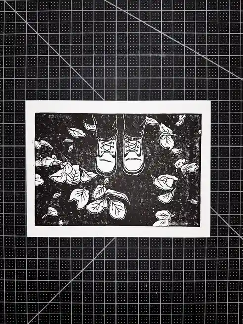 A black-and-white linocut print shows a pair of boots standing on a ground covered in scattered leaves, framed by a white…