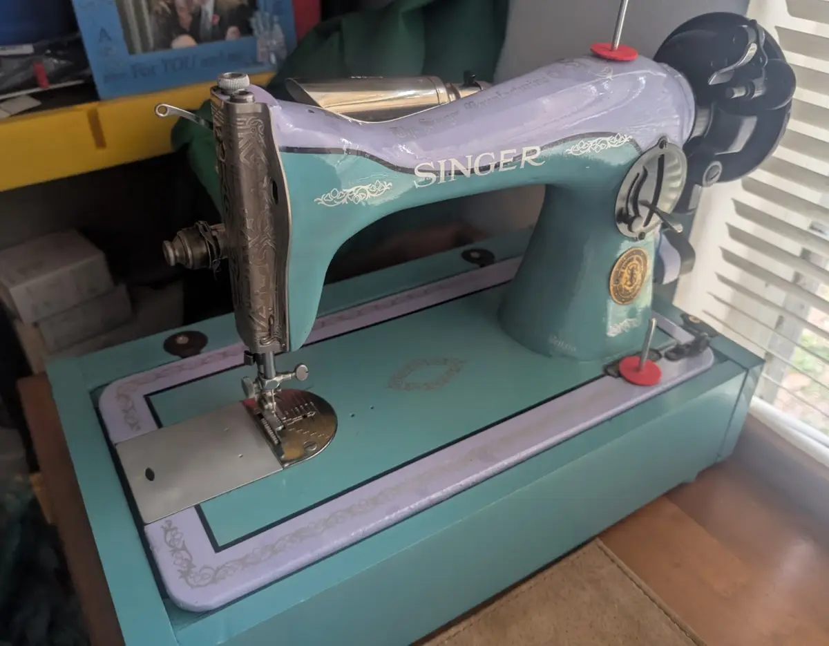 A vintage teal and lavender Singer sewing machine sits on a wooden surface, with ornate metal parts and a needle ready for…