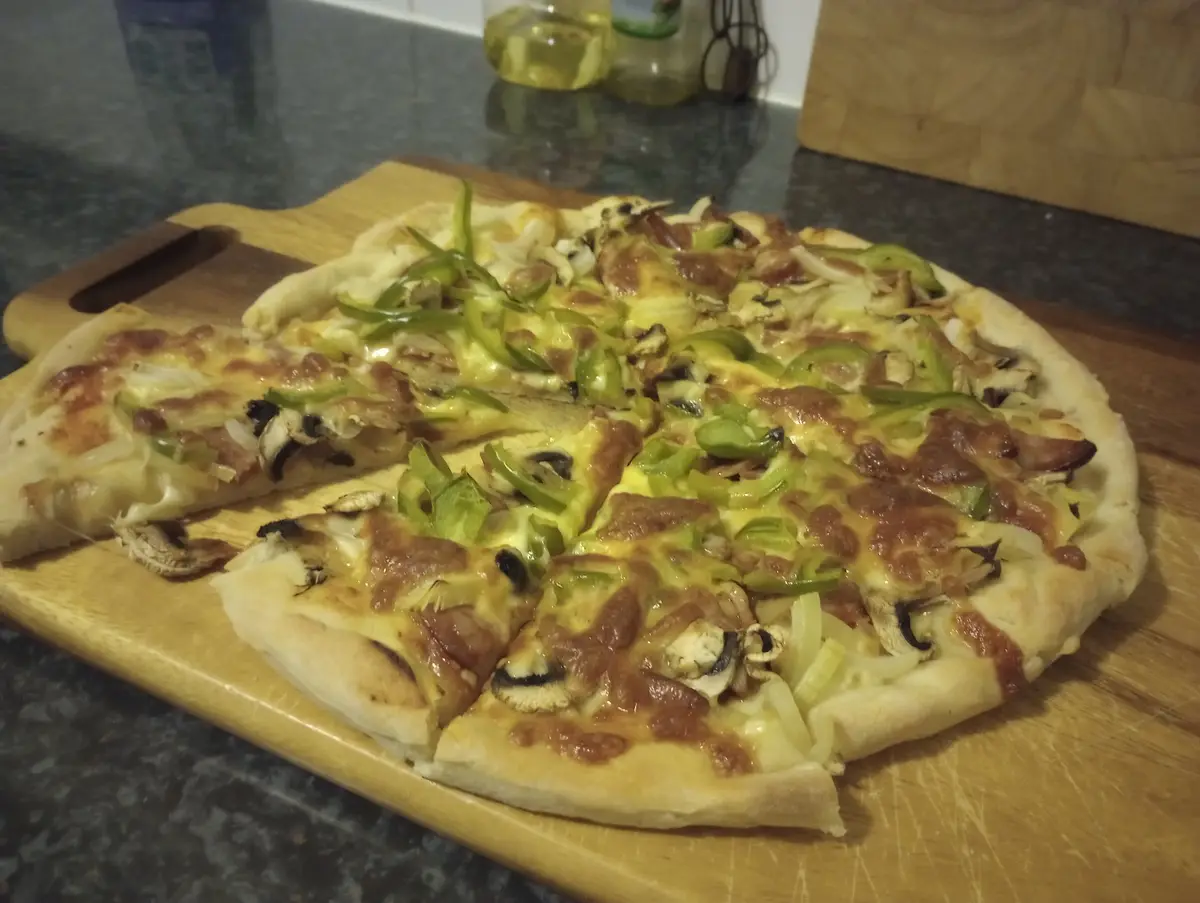 A homemade pizza with green peppers, mushrooms, onions, and cheese sits on a wooden cutting board with one slice removed…