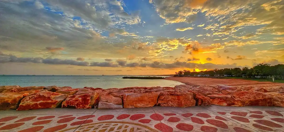 A scenic photo of a sunset over a calm ocean with a reddish-orange rocky barrier in the foreground and a patterned walkway…
