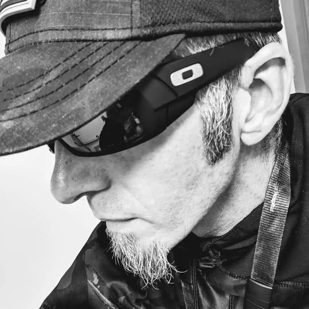 A black-and-white close-up of a man with gray hair and a goatee, wearing a cap and Oakley sunglasses, looking down with a…