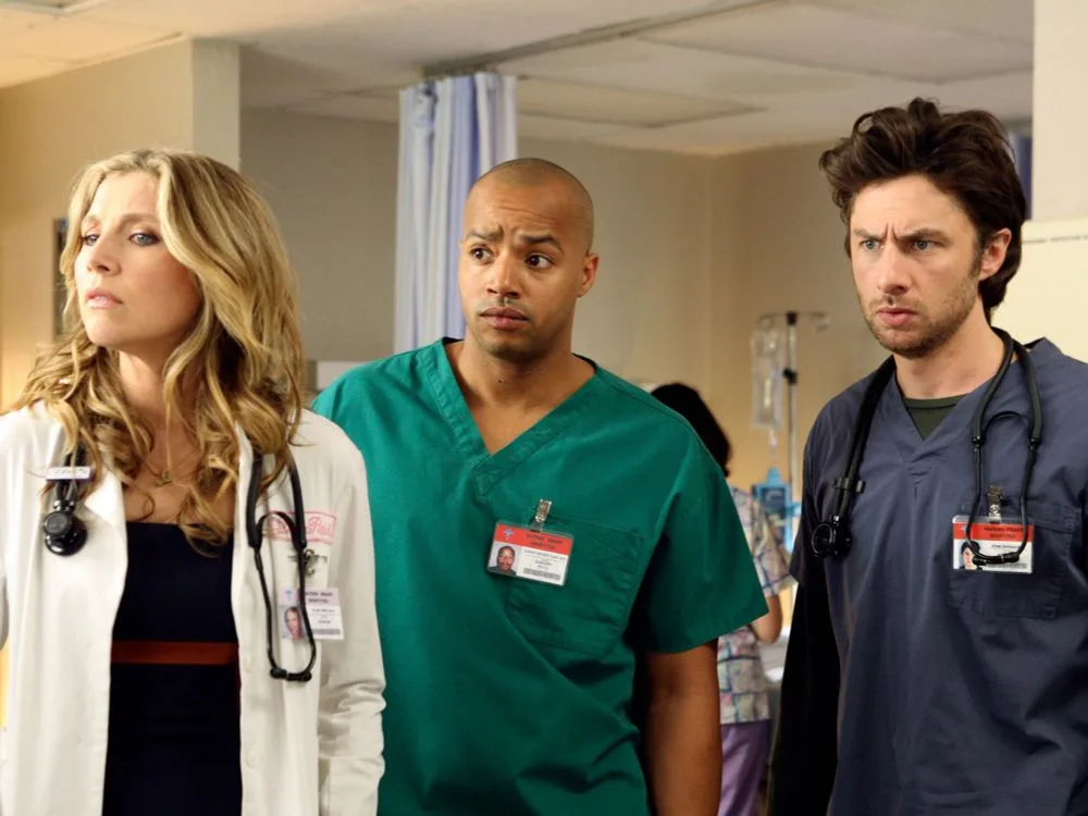 Three medical professionals in scrubs and lab coats stand in a hospital room, all looking confused and concerned. The woman…