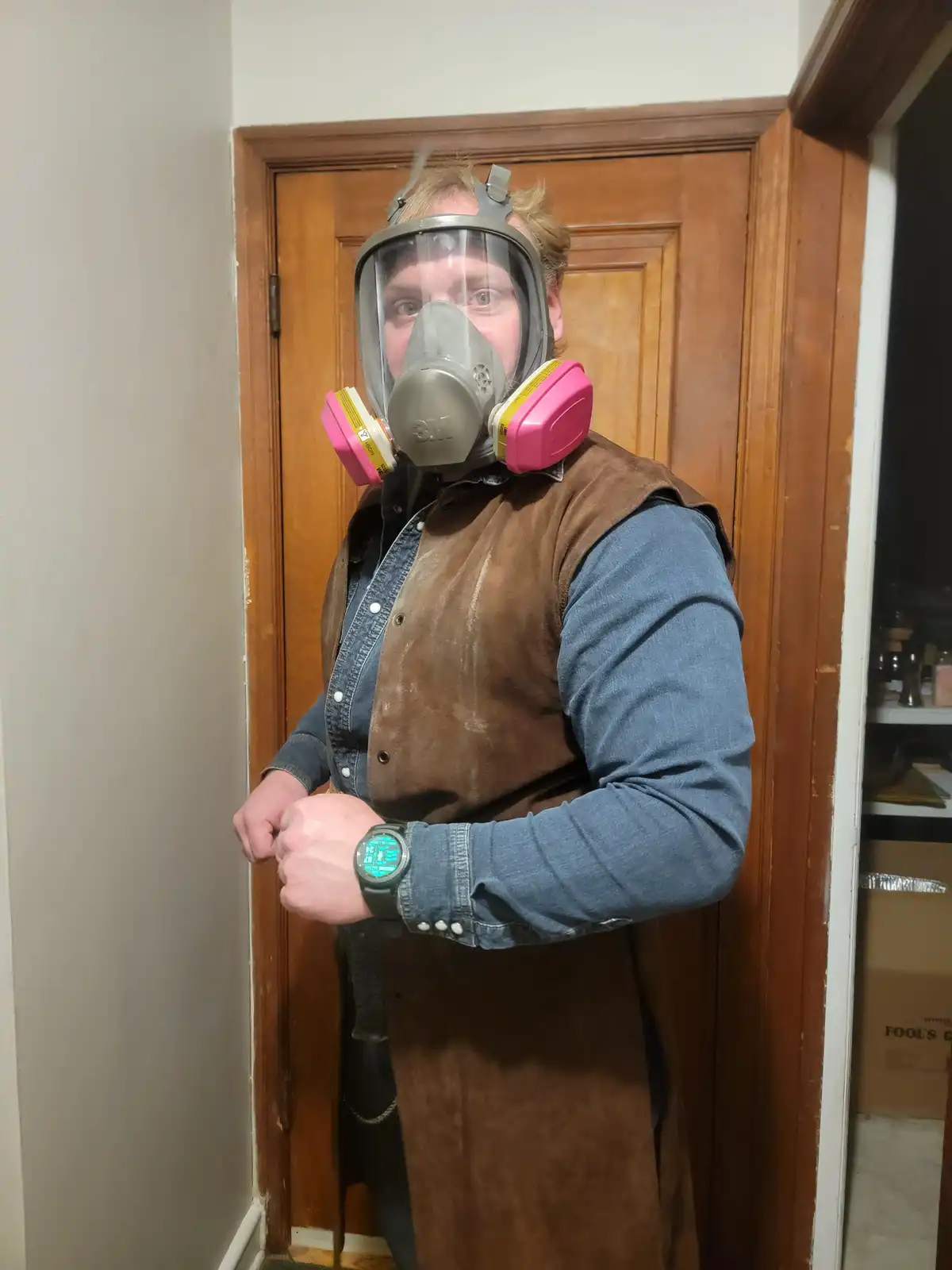 A man wearing a full-face 3M gas mask with pink filters stands in a doorway, looking at his glowing green digital watch. He…