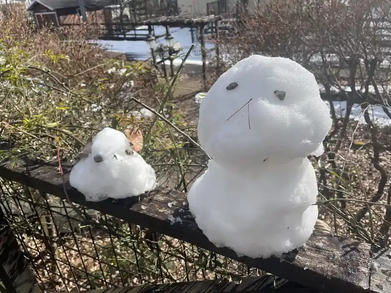 Two small snowmen sit on a wooden fence, one larger and one smaller, both with stone eyes and twig mouths, melting slightly…