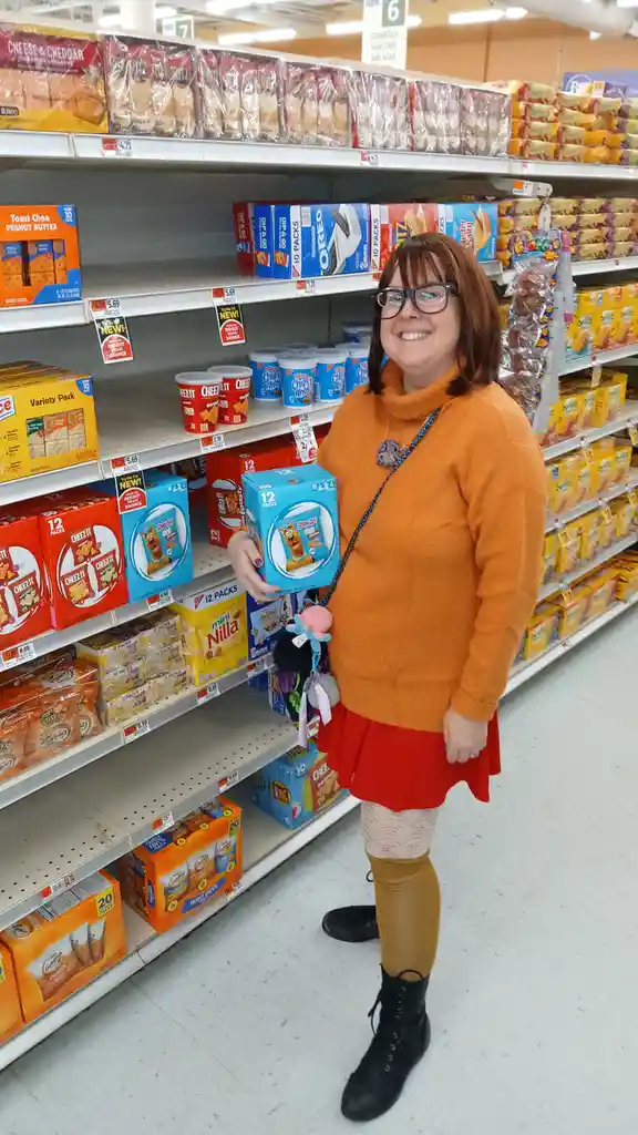 A woman in an orange sweater and red skirt stands in a grocery store aisle, smiling while holding a blue box of Scooby-Doo…