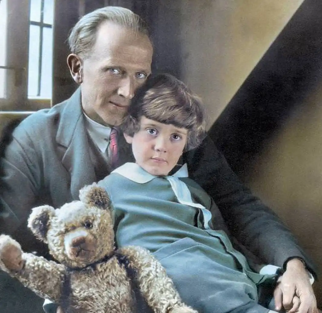 A vintage colorized photograph shows a man with slicked-back hair and a suit holding a young child in a green outfit. The…