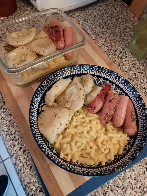 A plate and glass container hold a meal of macaroni and cheese, boiled pierogies, and grilled hot dogs, all seasoned with…