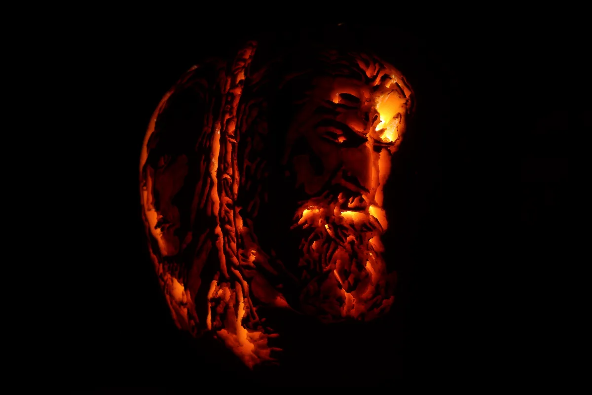 A close-up of a intricately carved pumpkin lantern glowing from within, depicting the face of Jesus Christ with detailed…