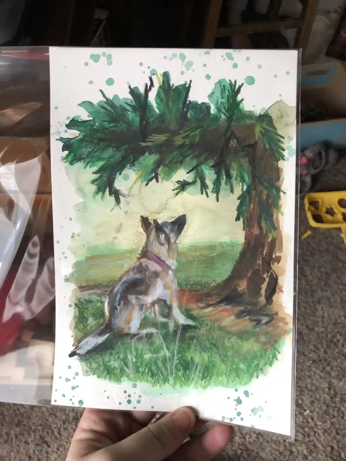 A hand holds a watercolor painting of a dog sitting under a pine tree, looking upward with an attentive expression. The…