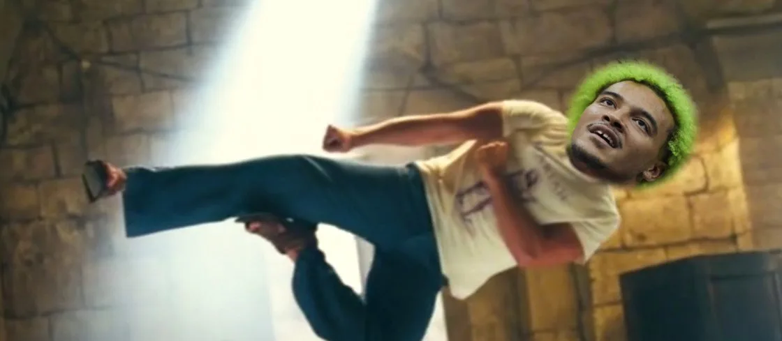 A man with bright green hair is shown mid-air in a dramatic pose, wearing a white t-shirt and blue pants, against a stone…