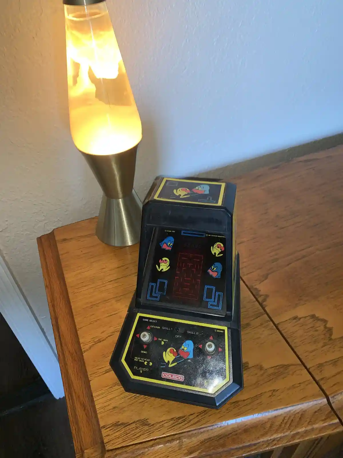 A vintage Coleco Pac-Man handheld game console sits on a wooden table next to a glowing lava lamp. The game's screen…