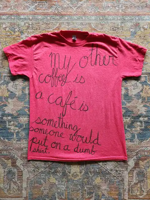 A bright pink t-shirt laid flat on a patterned rug, featuring black cursive text that reads 'my other coffee is a café is…