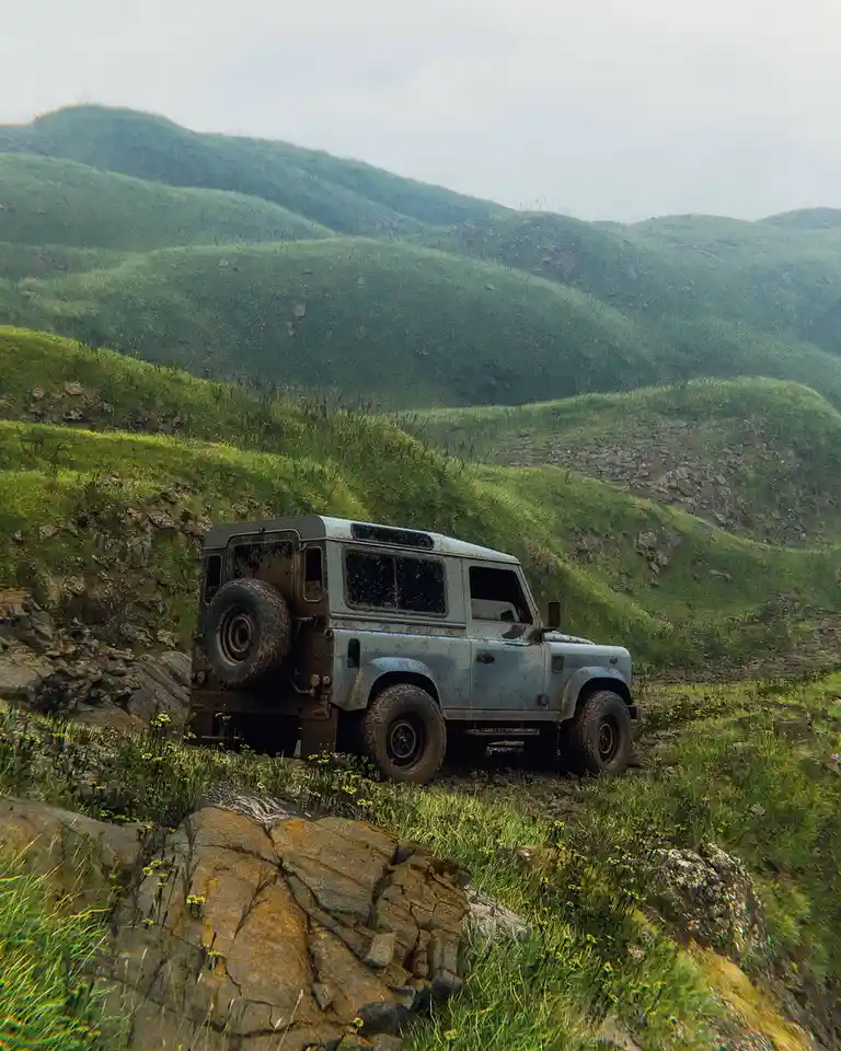 A weathered, light-blue Land Rover Defender is parked on a grassy, rocky hillside with rolling green hills in the…