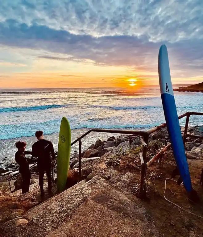 Two people in wetsuits stand on a rocky coastal path overlooking the ocean at sunset, holding surfboards. One green and one…