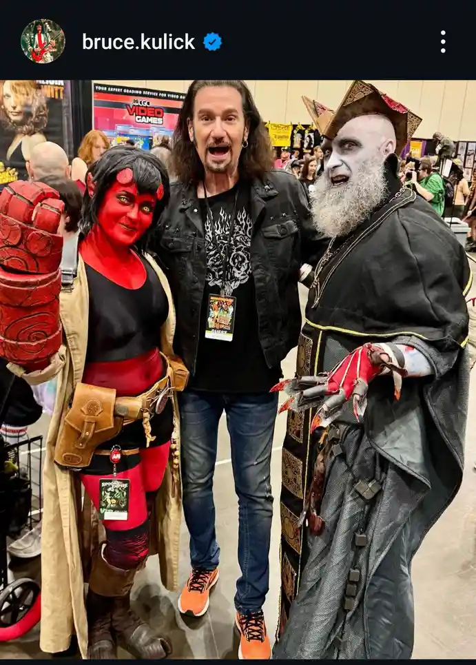 A man with long dark hair and a black jacket stands between two people in elaborate cosplay. On his left is a person dressed…