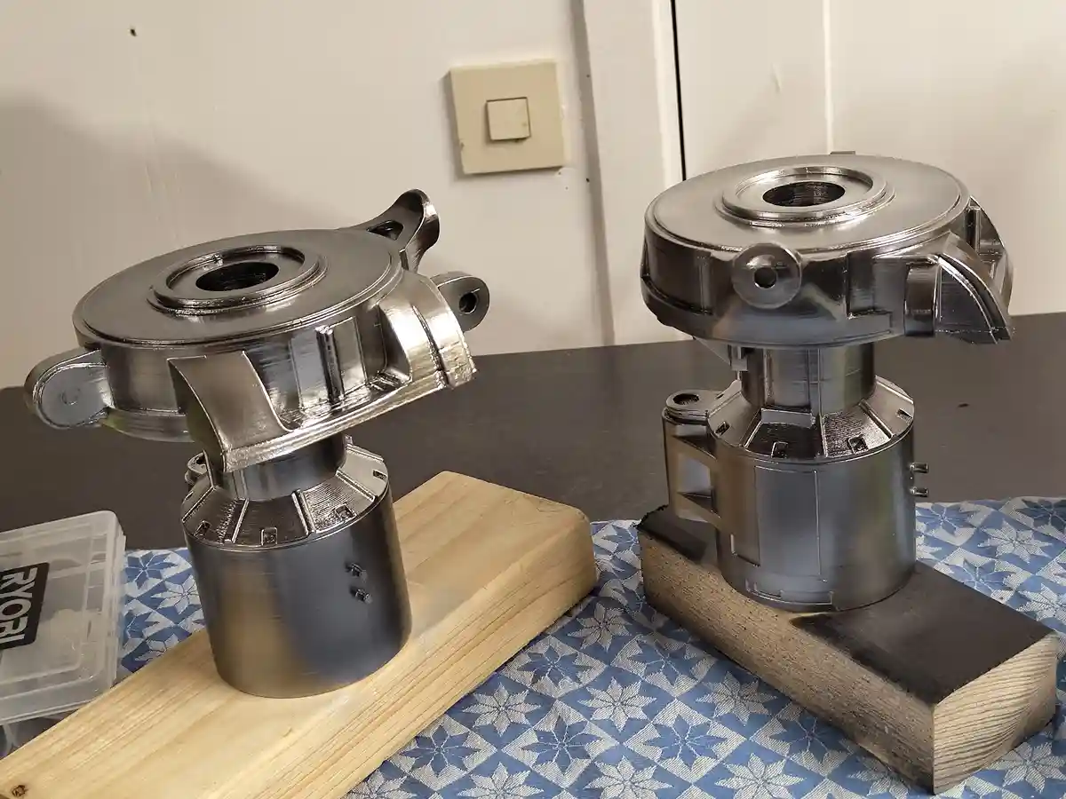 Two metallic, 3D-printed mechanical parts sit on wooden blocks on a blue patterned cloth; the left part has visible layer…