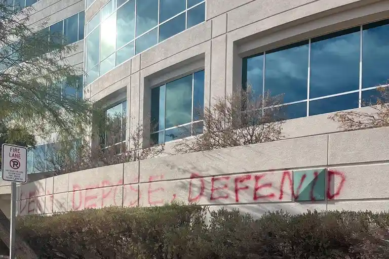 A photo of a modern office building with red graffiti on its lower wall that reads 'DEPOSE, DEFENID', which appears to be a…