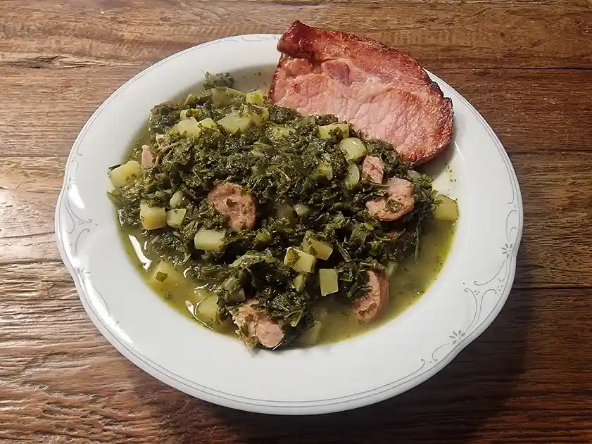 a white plate with decorative edges holds a serving of cooked collard greens mixed with diced potatoes and pieces of…