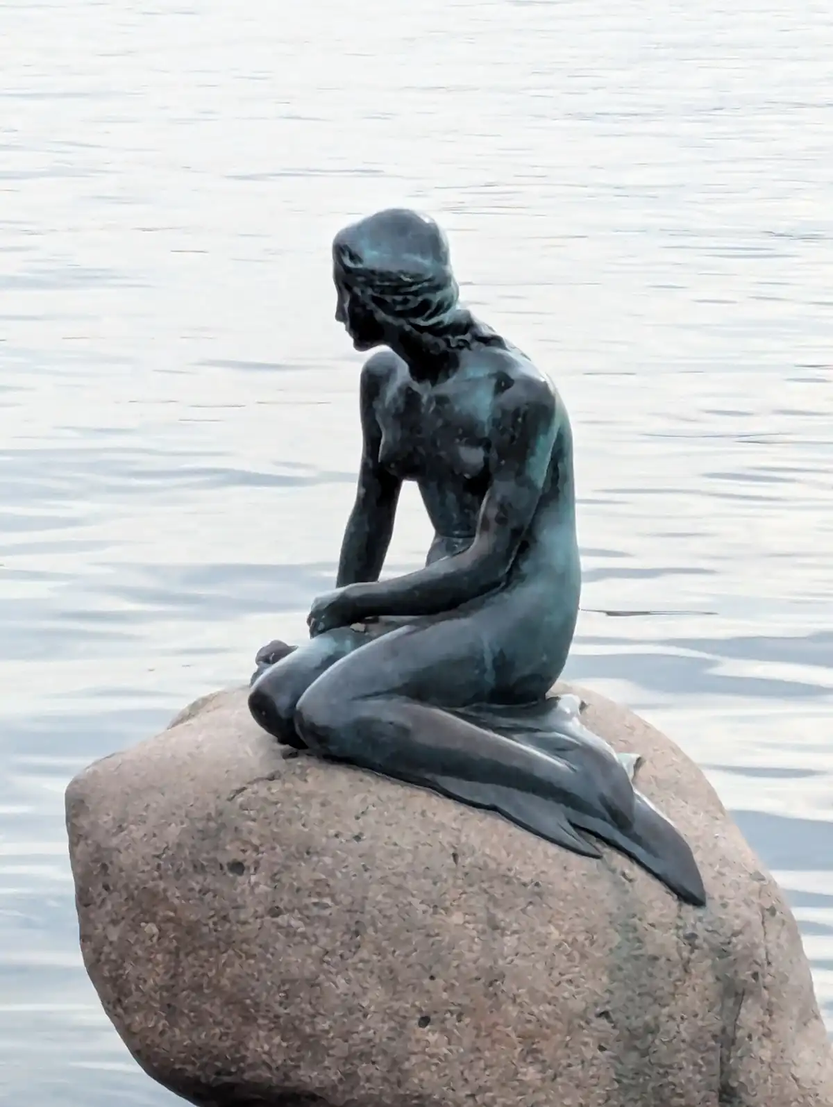 A bronze statue of a mermaid with a fish tail sits on a large rock partially submerged in water, looking downcast. The…