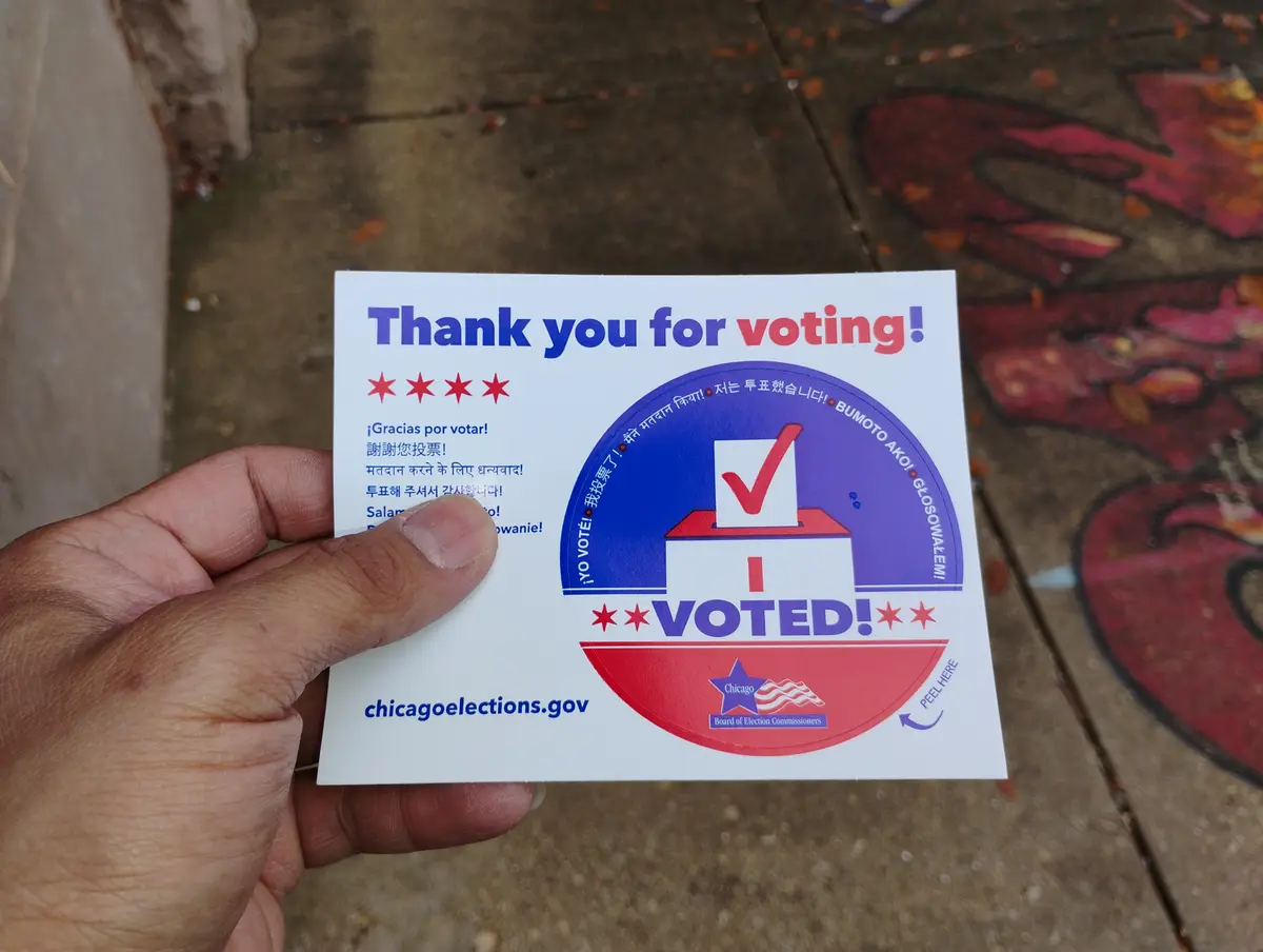 A hand holds a Chicago 'I Voted' sticker from chicagoelections.gov, featuring a red, white, and blue design with a ballot…