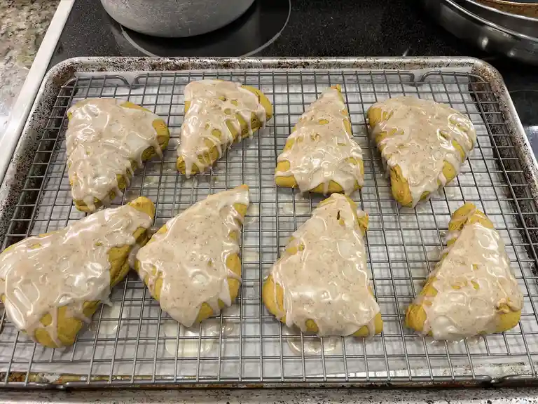 Eight golden-brown pumpkin scones, triangular in shape and drizzled with a white cinnamon glaze, are arranged on a metal…