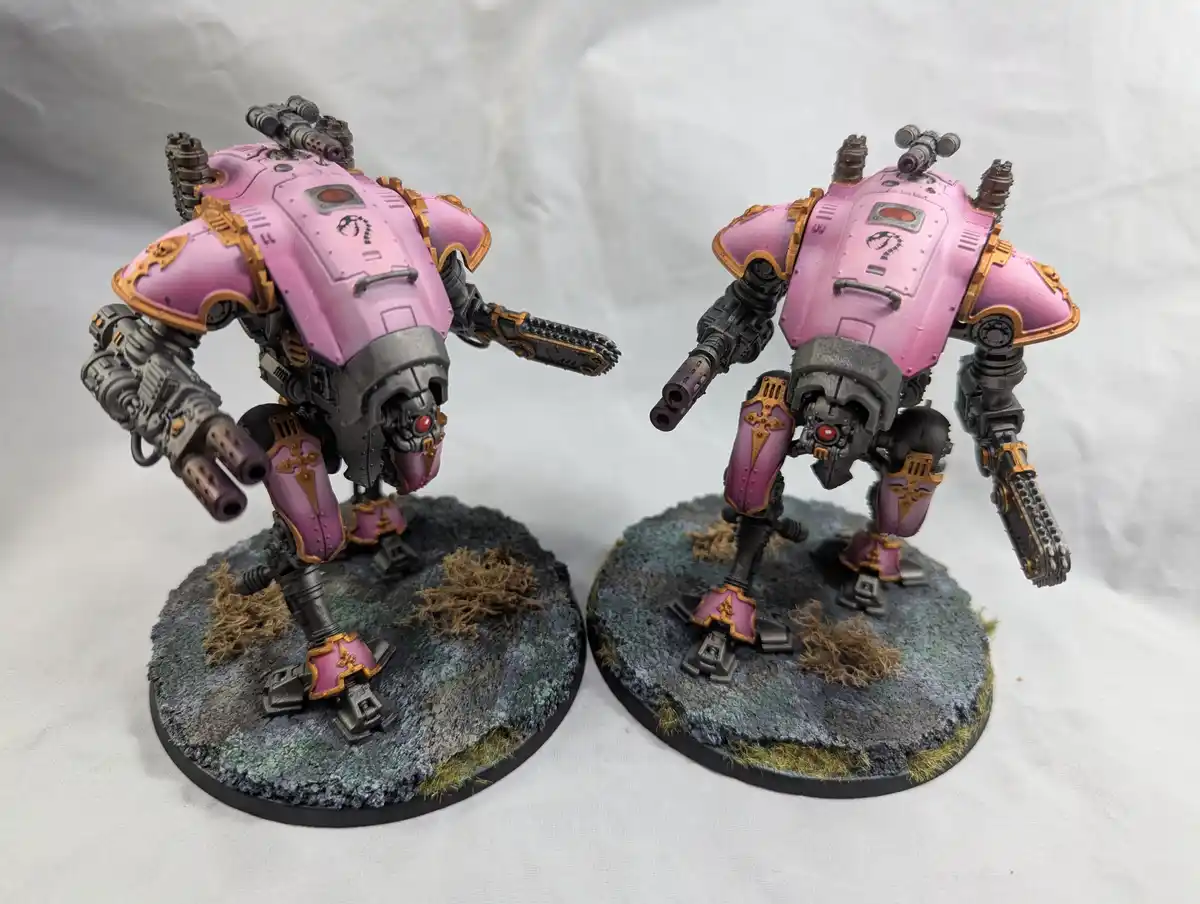 Two heavily armored, pink and gold painted war machines stand side by side on circular, textured bases with moss and dirt…