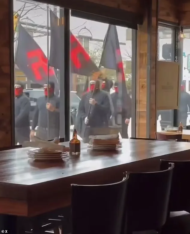 A view from inside a restaurant looking out a large window at a group of people in dark clothing and red face coverings…